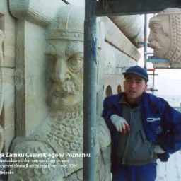 Pracownik w niebieskiej kurtce i czapce, wsparty o rusztowanie, na tle kamiennej płaskorzeźby rycerza i fragmentu elewacji wieży zegarowej podczas renowacji Zamku Cesarskiego w Poznaniu.