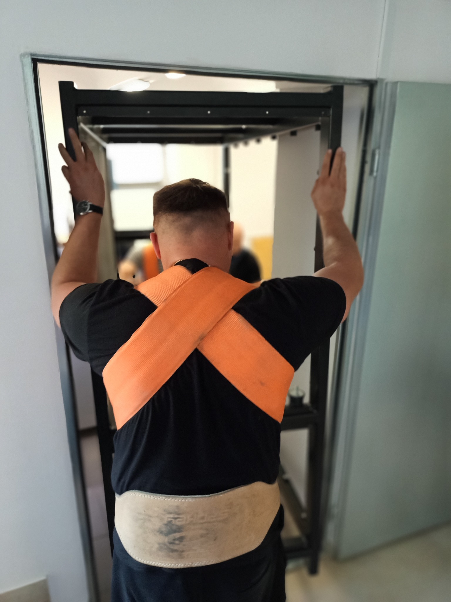Mężczyzna w czarnej koszulce i pomarańczowych pasach transportowych, tyłem do kamery, przenosi metalowy regał przez drzwi, widoczny pasek ochronny na lędźwiach.