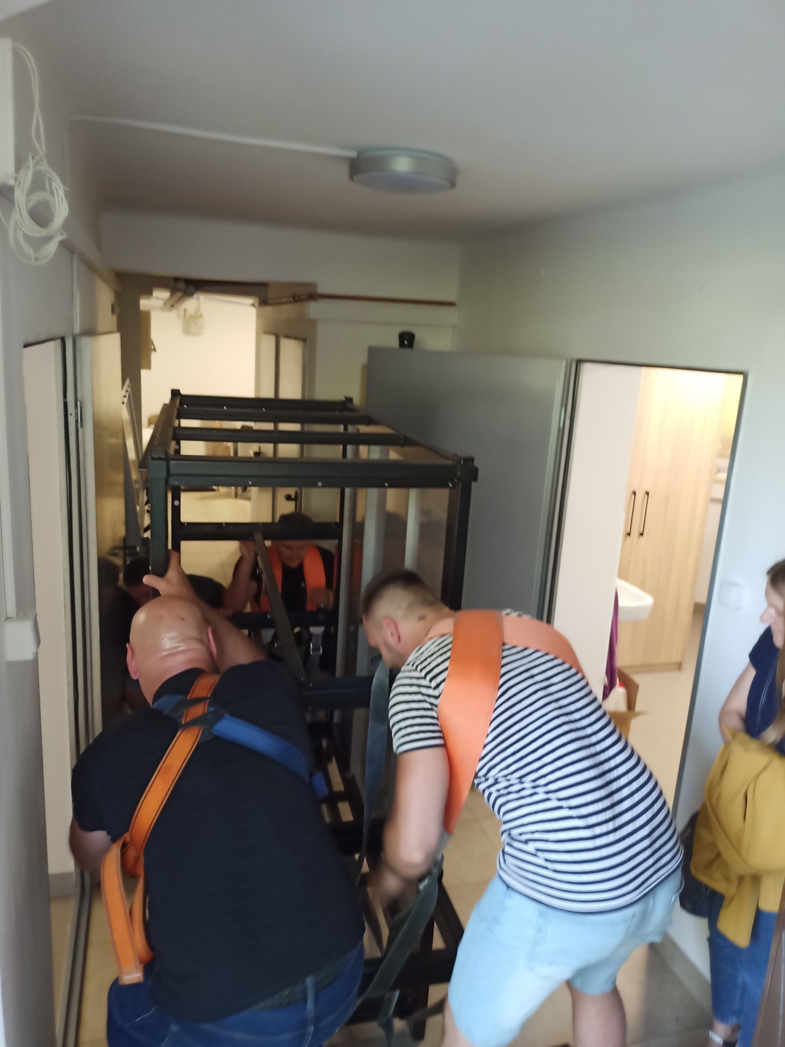 Dwóch mężczyzn w uprzężach transportuje metalową konstrukcję przez wąski korytarz, trzecia osoba obserwuje.