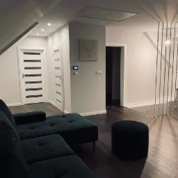 Nowoczesne wnętrze poddasza w Niedźwiedziu z ciemną podłogą, szarą ścianą z dekoracją jelenia, białymi drzwiami z przeszkleniami i ciemnozielonym narożnikiem oraz metalową balustradą schodową.