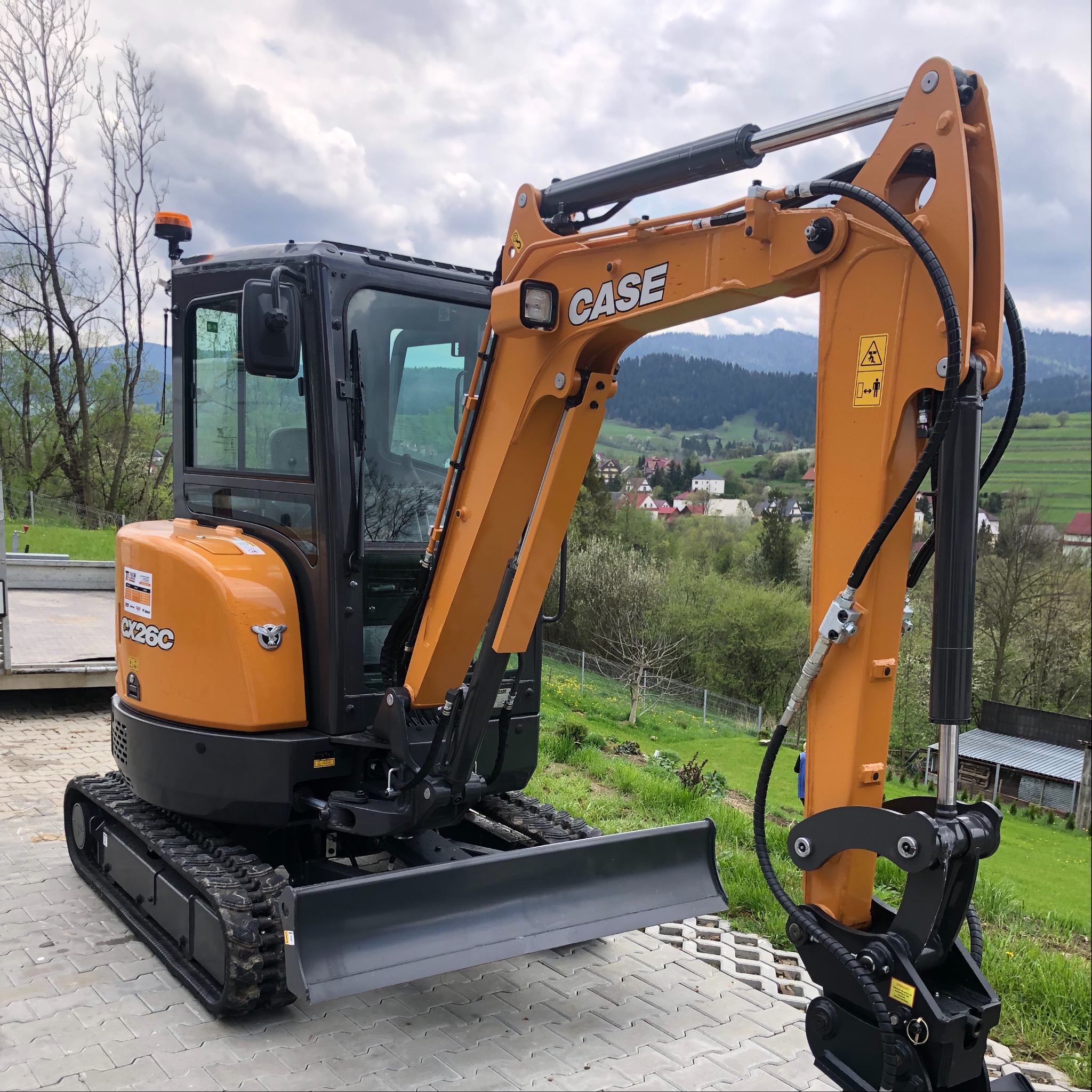 Pomarańczowo-czarna mini koparka Case CX26C na betonowej kostce, z widocznym osprzętem roboczym i malowniczym krajobrazem w tle.