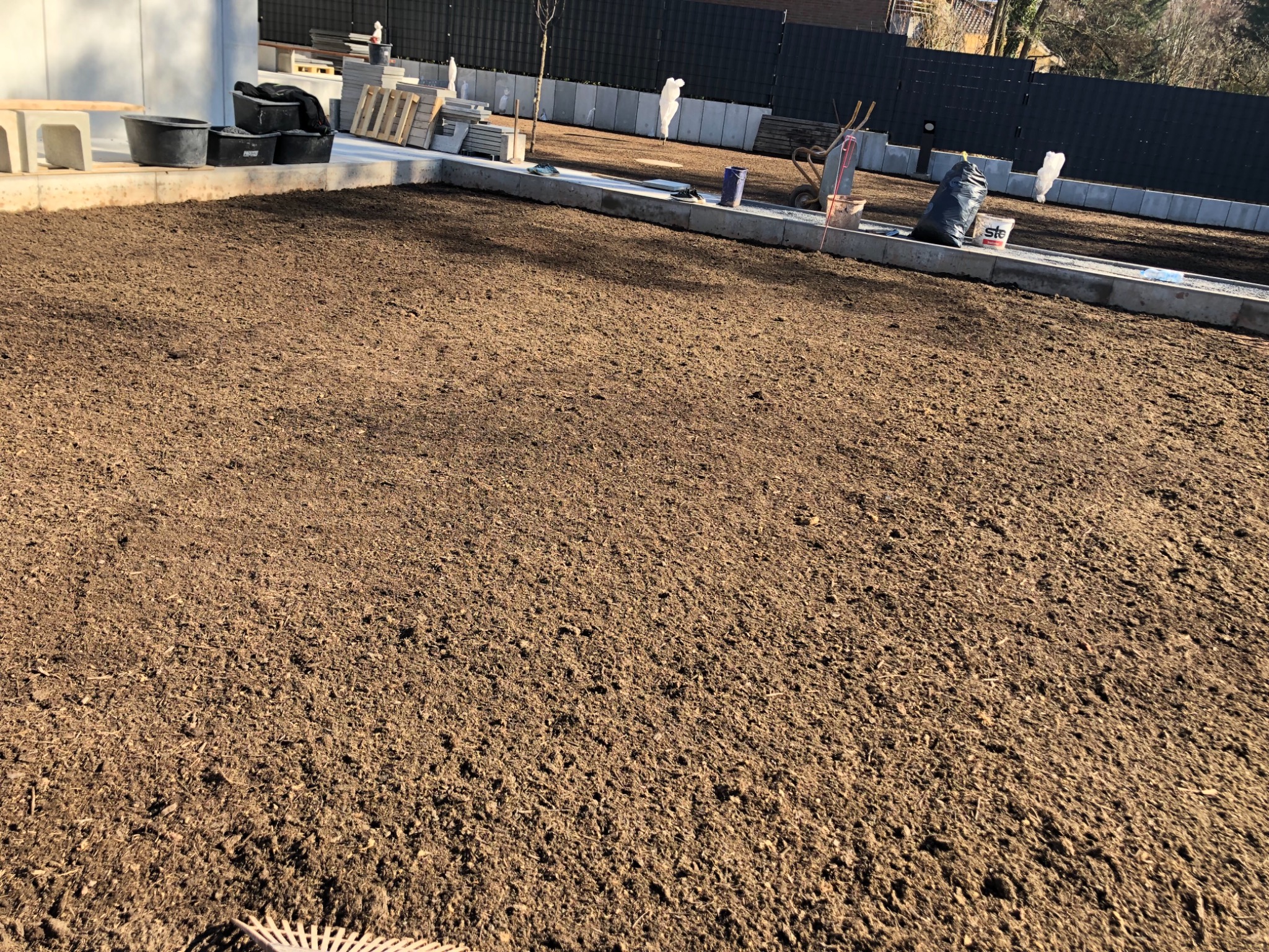 Przygotowany teren pod budowę, wyrównana ziemia otoczona betonową obwódką, widoczne narzędzia i materiały budowlane w tle.