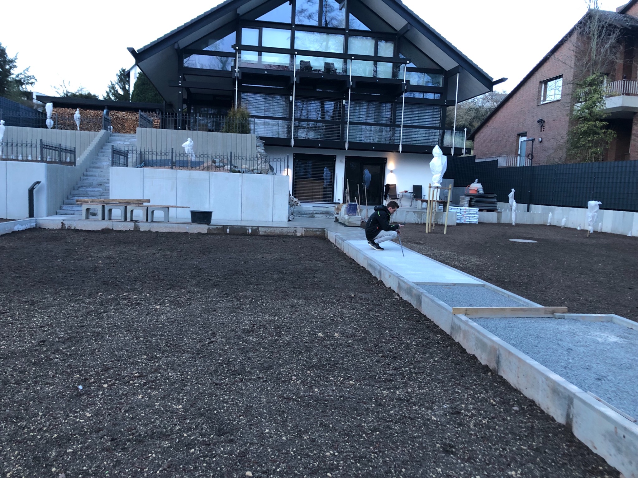 Pracownik w trakcie wyznaczania poziomu nowej ścieżki z obrzeżami betonowymi na posesji z nowoczesnym domem i schodami na podwyższeniu.