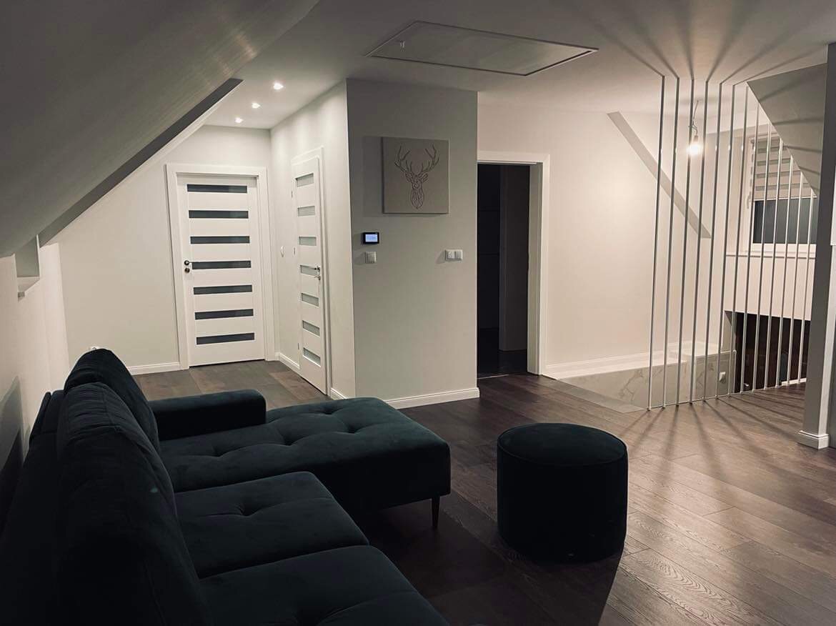 Nowoczesne wnętrze poddasza w Niedźwiedziu z ciemną podłogą, szarą ścianą z dekoracją jelenia, białymi drzwiami z przeszkleniami i ciemnozielonym narożnikiem oraz metalową balustradą schodową.