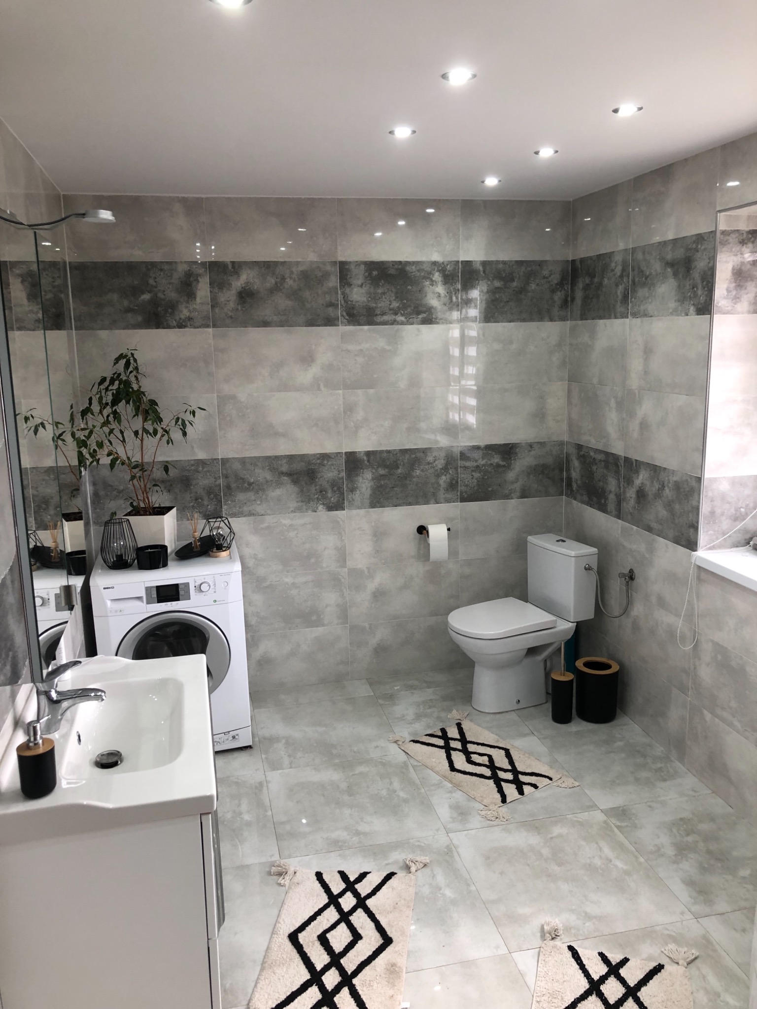 Jasna łazienka z szaro-białymi płytkami imitującymi marmur, białą umywalką z szafką, toaletą, pralką i dekoracyjnymi dywanikami z geometrycznym wzorem.