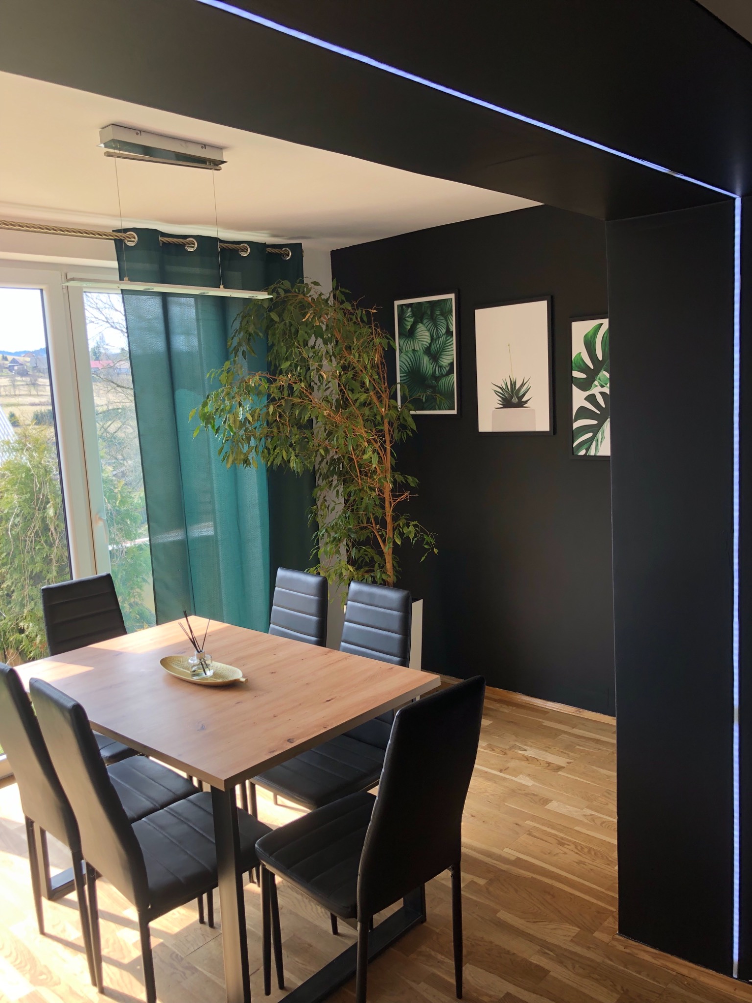 Nowoczesna jadalnia z drewnianym stołem, czarnymi krzesłami, parkietem i czarną ścianą z oświetleniem LED oraz dekoracjami roślinnymi.