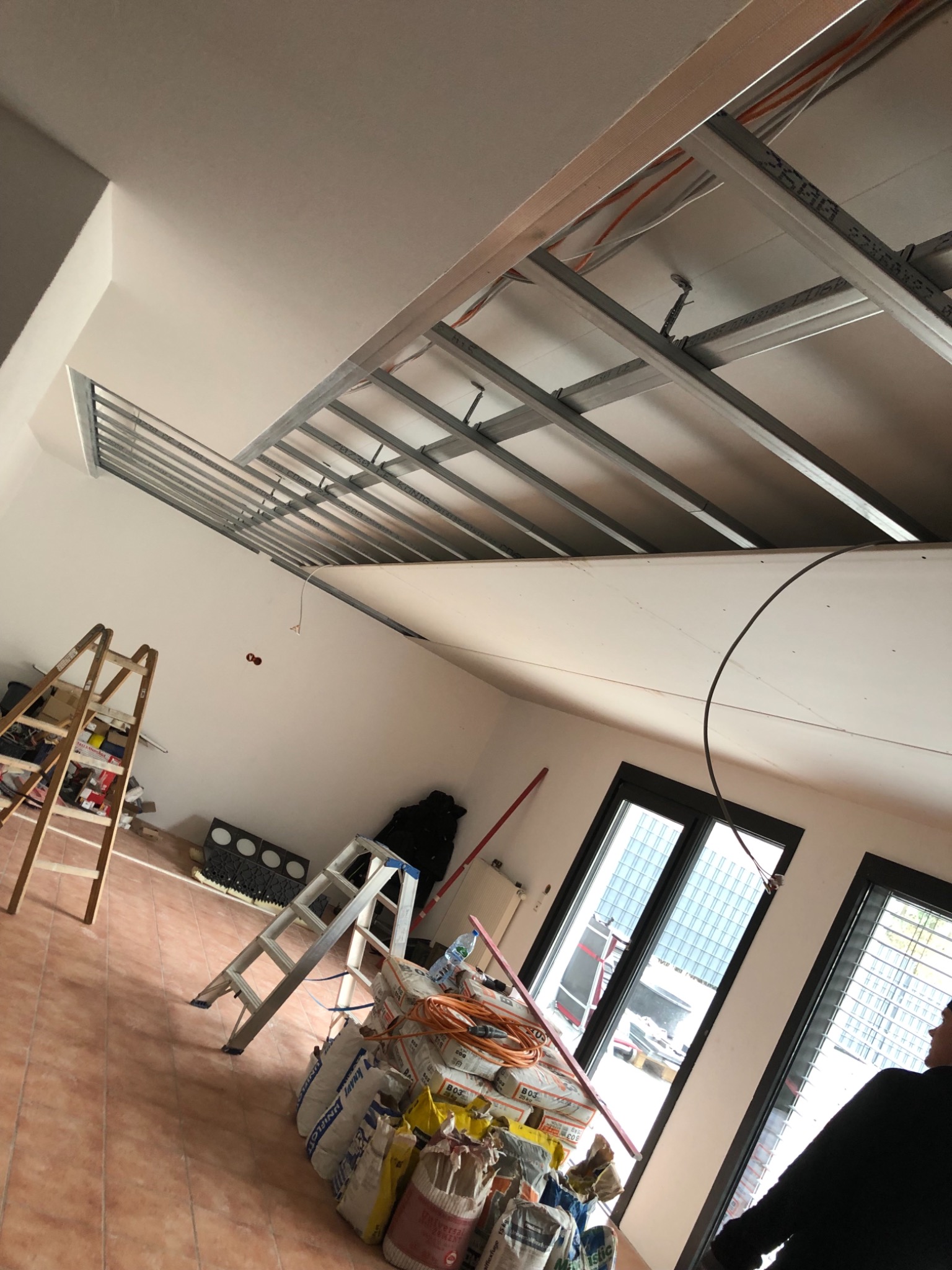 Wnętrze w trakcie remontu: montaż metalowej konstrukcji sufitu podwieszanego, widoczne worki z zaprawą, drabiny i okna.