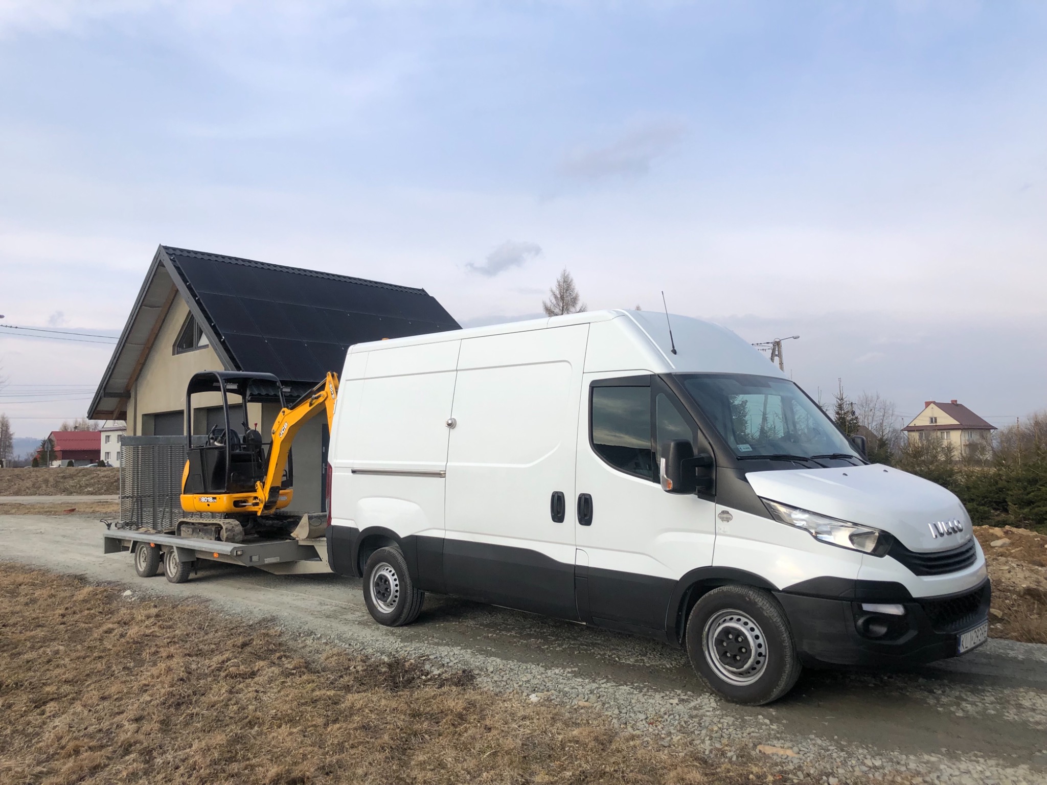 Biały furgon Iveco z czarnym pasem holuje na przyczepie żółtą mini koparkę JCB 8018 CTS na tle domu z czarnym dachem i pochmurnego nieba.