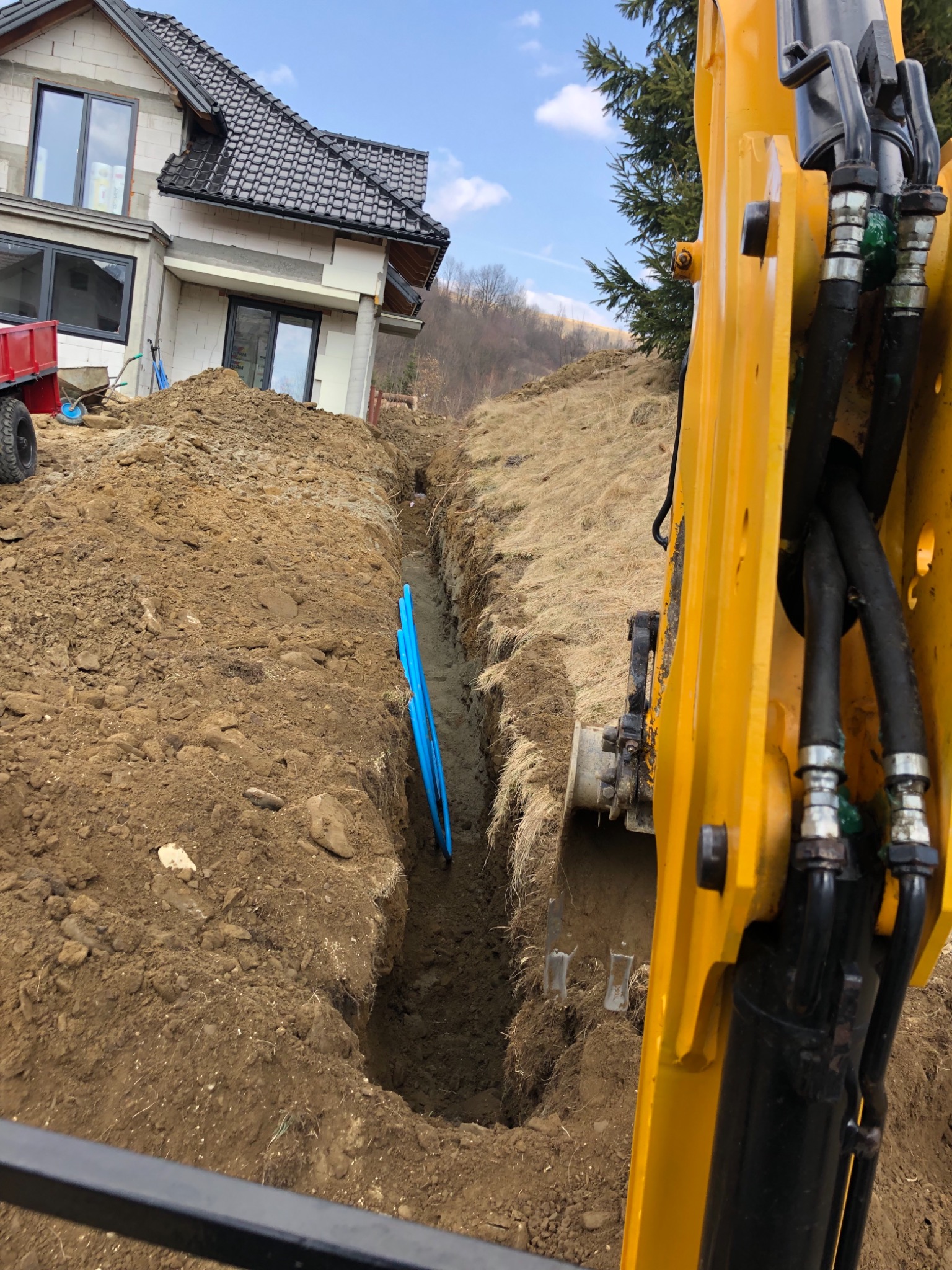 Głęboki wykop w ziemi, z widocznymi niebieskimi rurami, obok żółta koparka, w tle budynek w trakcie budowy i zalesione wzgórze.