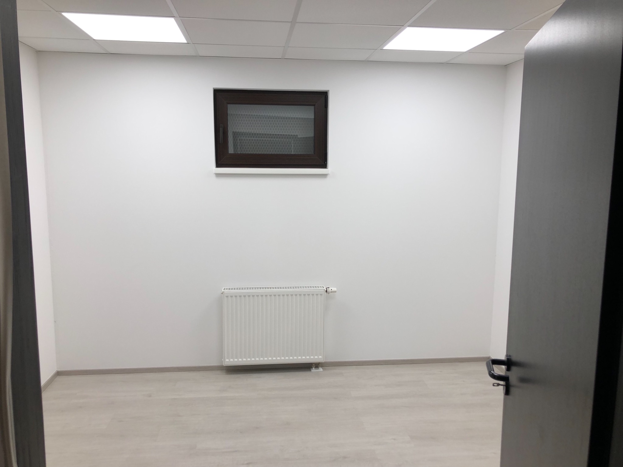 Puste, białe pomieszczenie biurowe z grzejnikiem, oknem z kratą i podwieszanym sufitem, widok zza otwartych drzwi.
