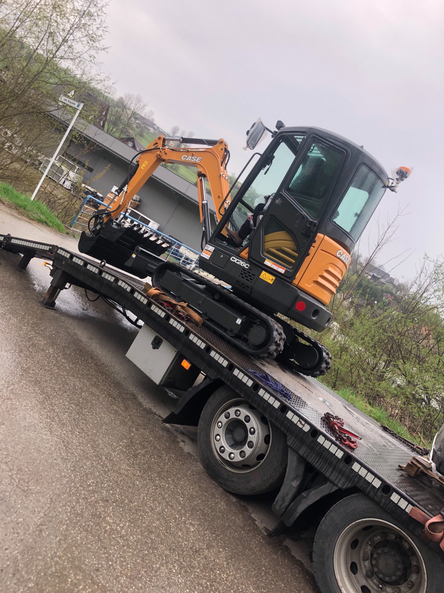 Pomarańczowo-czarna minikoparka Case CX26C na lawecie transportowej, widok z boku na tle zabudowań i zieleni w pochmurny dzień.