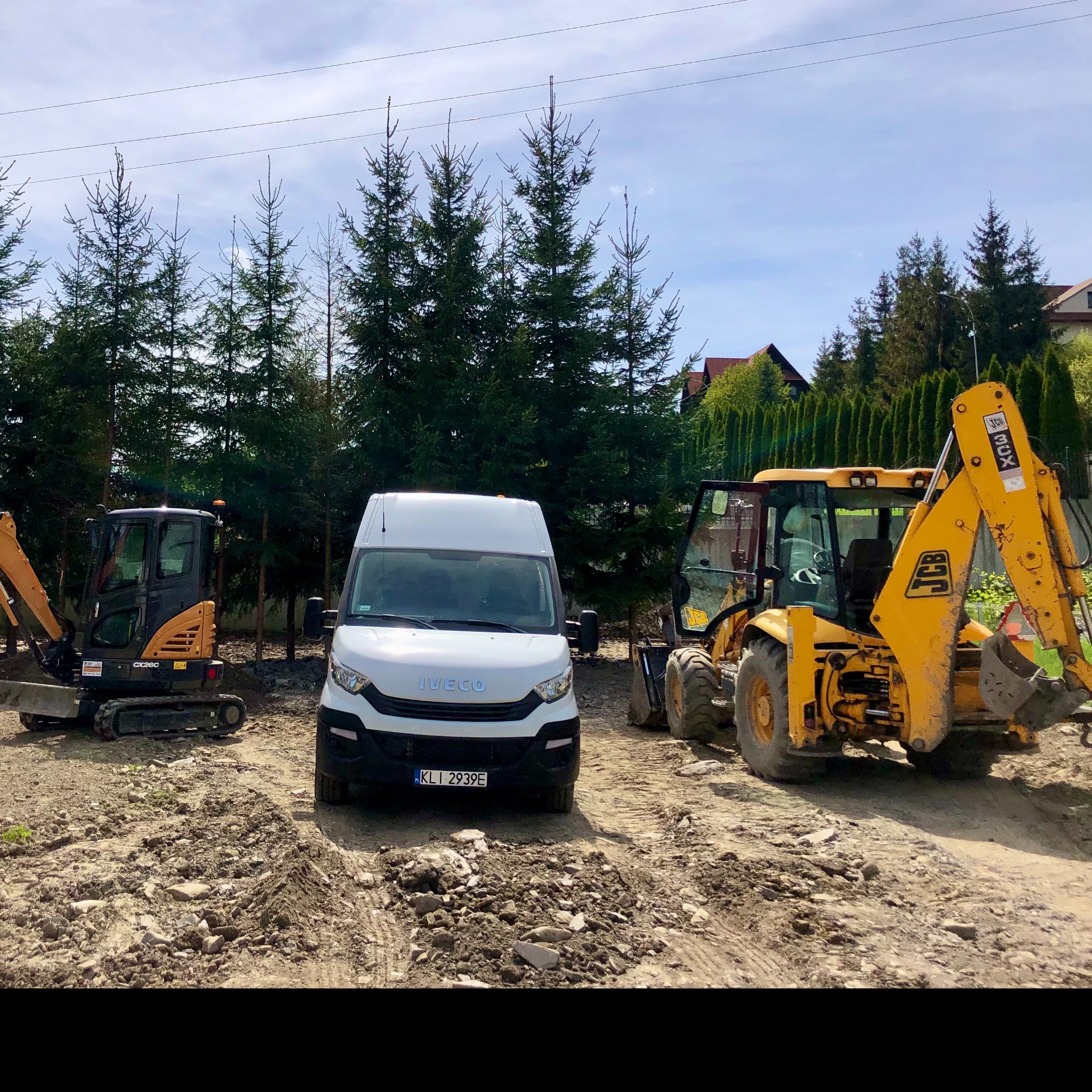 Żółta koparko-ładowarka JCB 3CX, mała koparka i biały samochód dostawczy Iveco na placu budowy z ziemią i gruzem, w tle rząd drzew iglastych i domy.