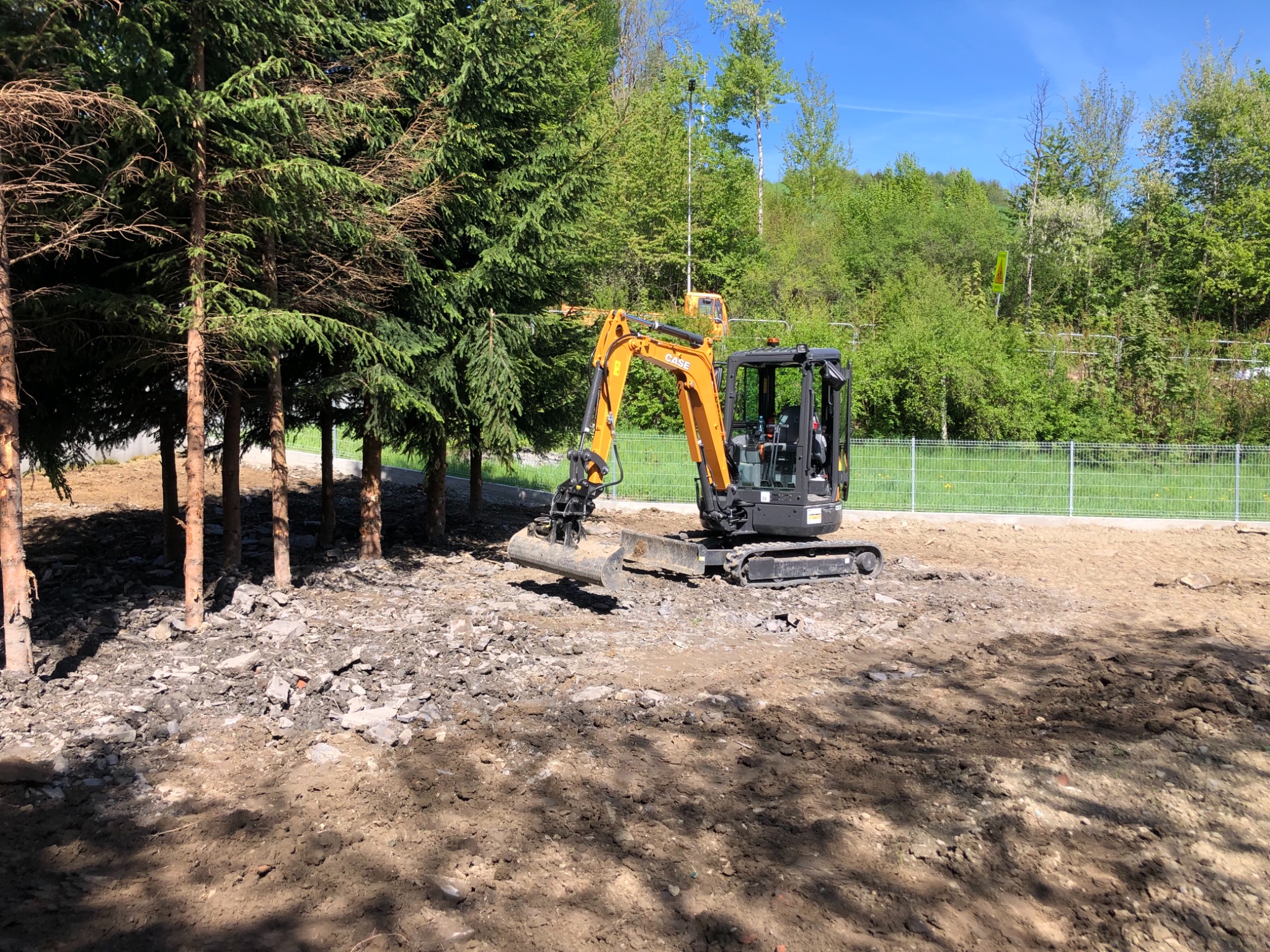 Mała pomarańczowa koparka CASE na placu budowy, przygotowująca teren pod przyszłą inwestycję, widoczne drzewa i ogrodzenie.