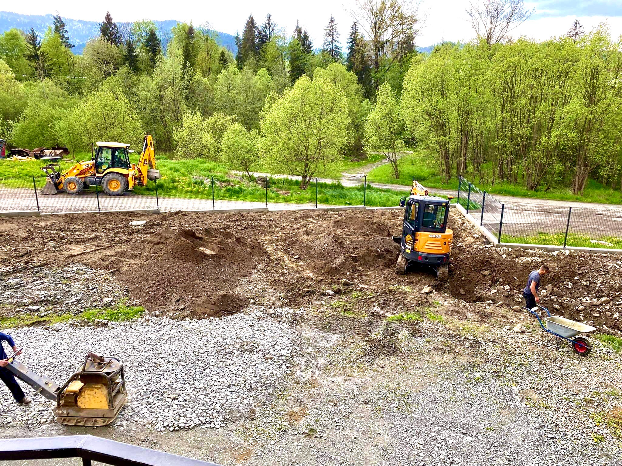 Żółta koparka i mini koparka podczas prac ziemnych na działce z widokiem na las, ubijarka wibracyjna i pracownik z taczką w pobliżu.