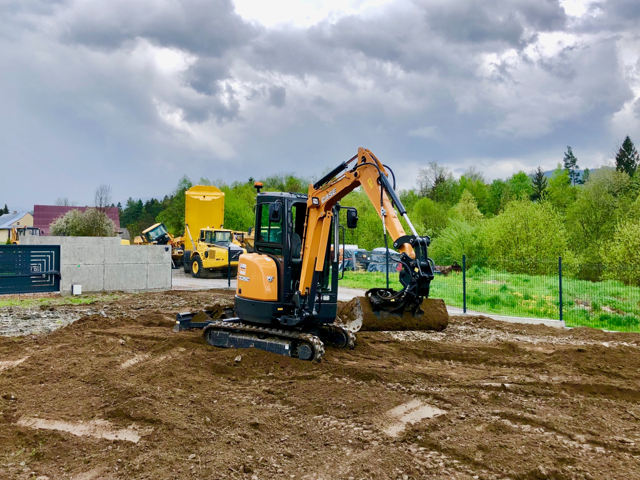 Pomarańczowo-czarna minikoparka Case CX26C na placu budowy, w tle żółte maszyny budowlane i zieleń drzew, pochmurne niebo.