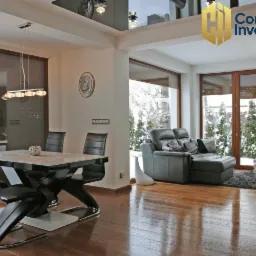 Jasne, przestronne wnętrze domu z parkietem, widok na ogród zimą przez duże okna, nowoczesny stół z krzesłami i skórzana sofa. W górnym rogu logo firmy Commercial Investments.