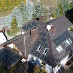 Widok z lotu ptaka na dom z brązowym dachem, na którym zamontowane są panele słoneczne; dodatkowe panele leżą na ziemi obok zaparkowanego białego vana i czarnego SUV-a.