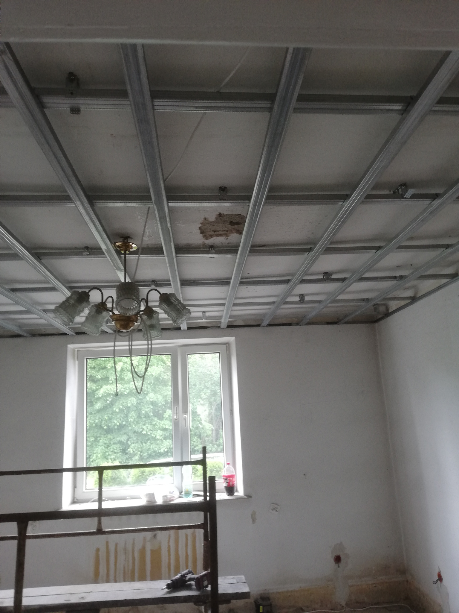 Montaż stelaża sufitu podwieszanego w pokoju z oknem, widoczny stary żyrandol i narzędzia na rusztowaniu.