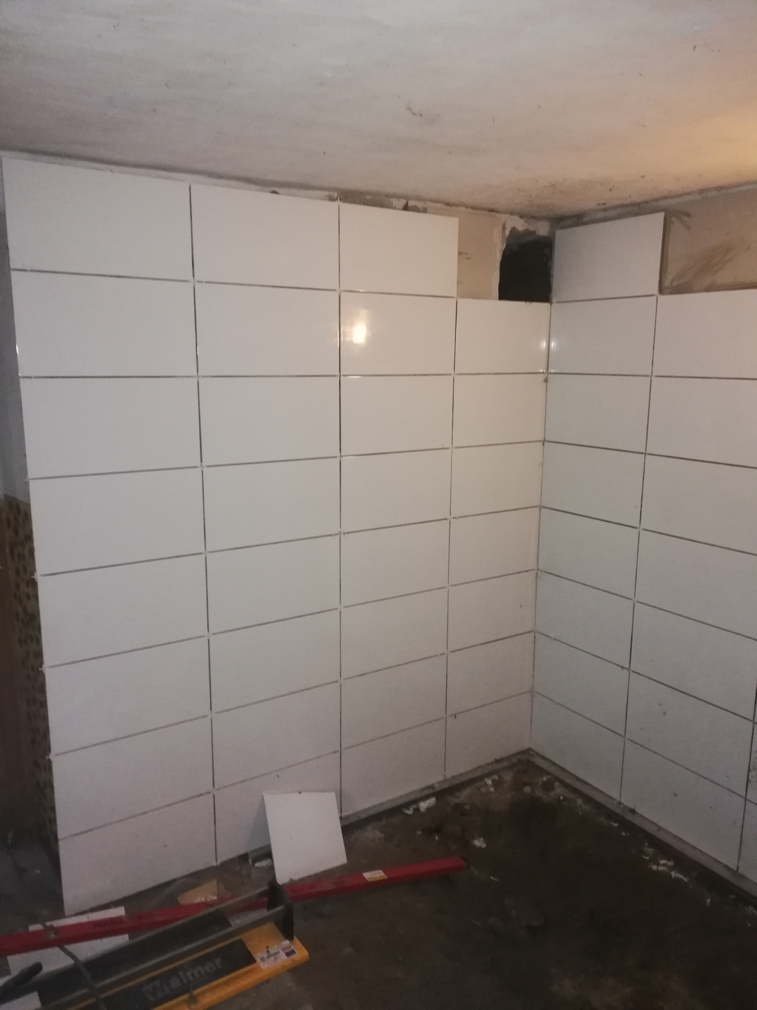 Ściany wyłożone białymi, prostokątnymi płytkami, widoczny fragment surowego sufitu i podłogi w trakcie prac wykończeniowych, z narzędziami do cięcia płytek na pierwszym planie.