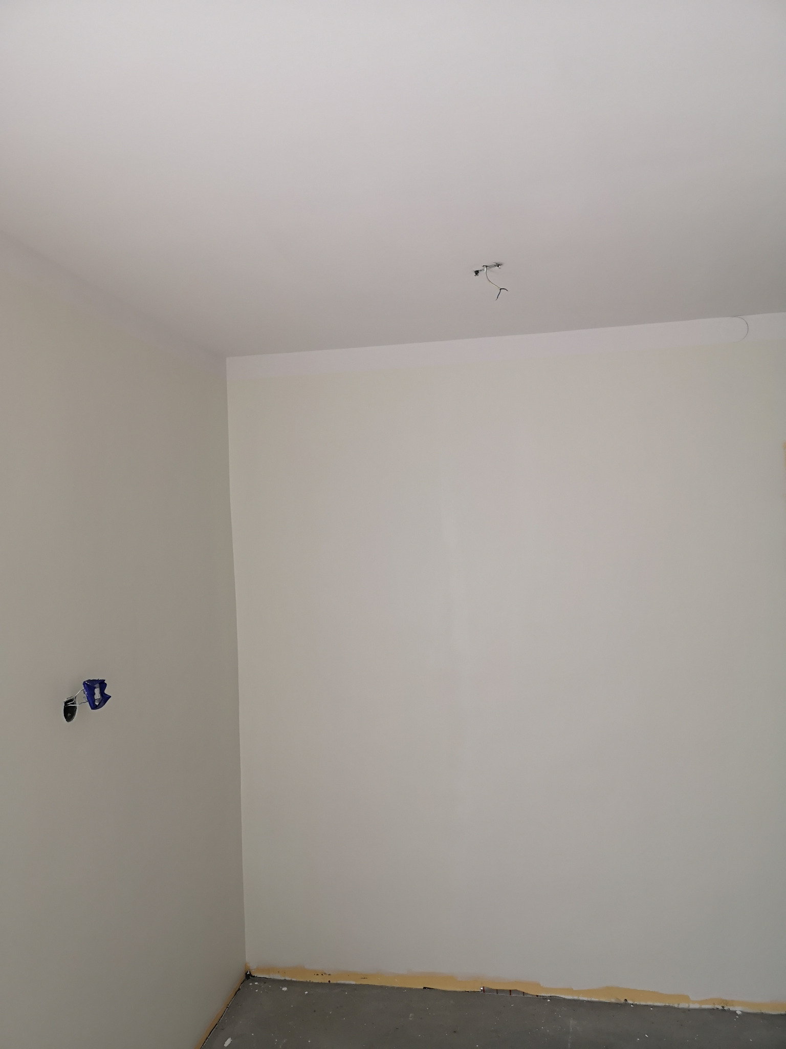 Pomalowane na jasny odcień pomieszczenie w trakcie wykańczania, widoczne wystające kable w suficie i niezabezpieczone gniazdko elektryczne na ścianie.