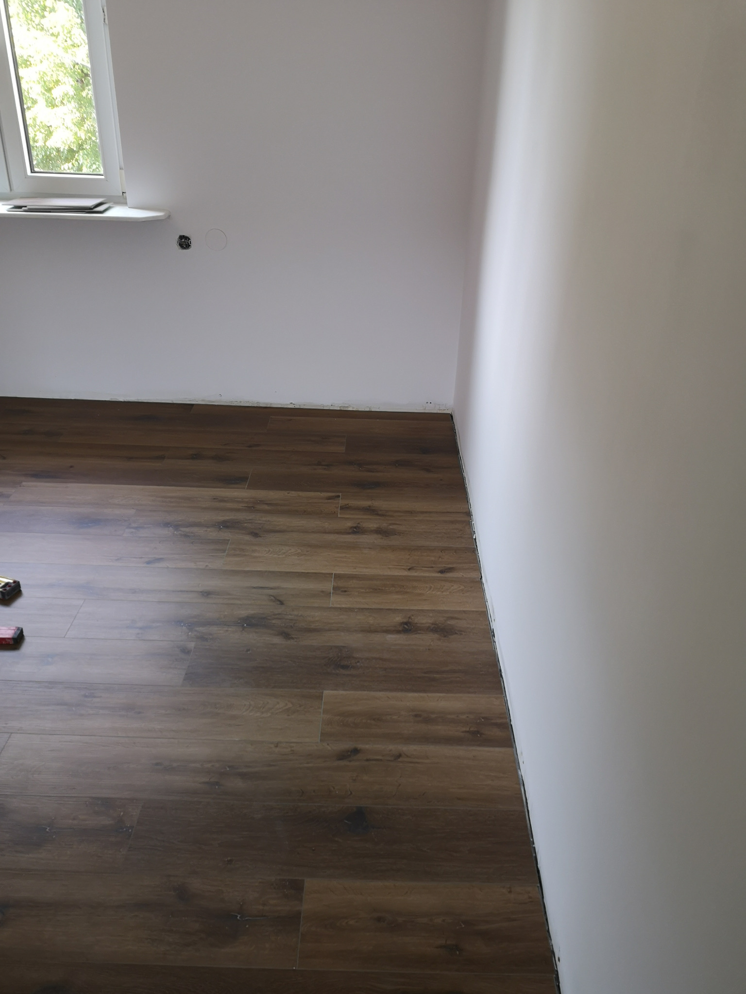 Ułożona, brązowa podłoga panelowa w rogu pokoju z białymi ścianami i oknem.