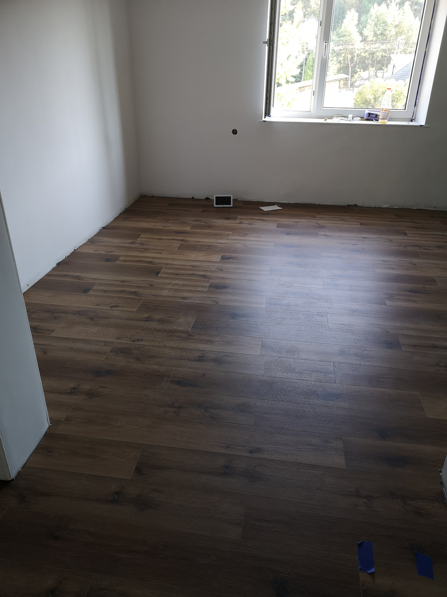 Ułożony parkiet w pokoju z oknem, widoczne niebieskie paski taśmy malarskiej na podłodze i drobne niedociągnięcia przy ścianach.