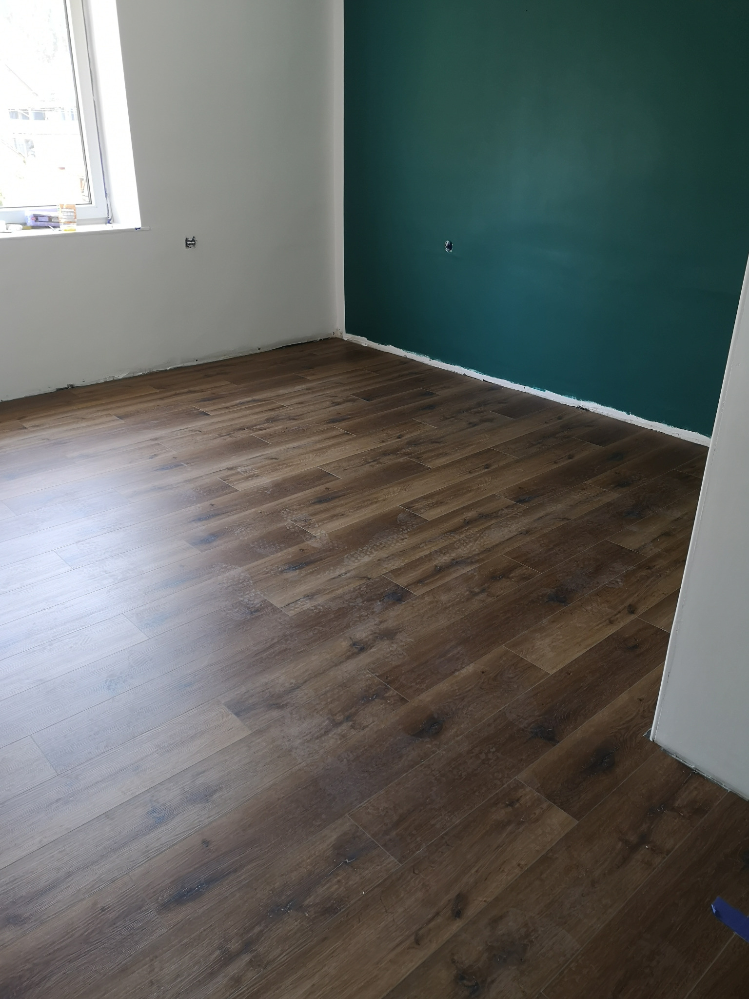 Pokój po remoncie: świeżo ułożony parkiet o ciemnym, drewnopodobnym wzorze, kontrastujący z pomalowaną na ciemnozielono ścianą i białą ścianą przy oknie, widoczne ślady po malowaniu przy podłodze.