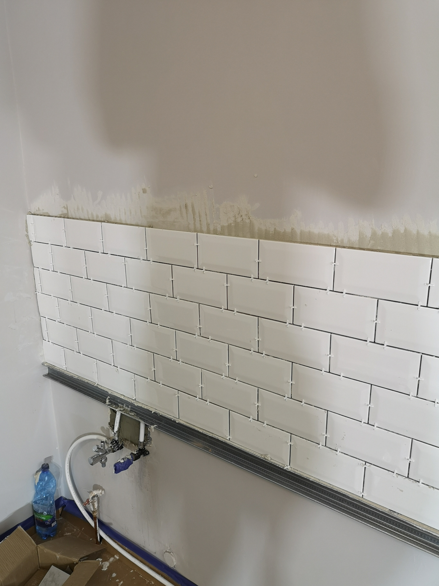 Białe, prostokątne płytki ułożone na ścianie z użyciem krzyżyków dystansowych, widoczna metalowa listwa podtrzymująca, ślady zaprawy na ścianie powyżej, instalacja wodna w tle.