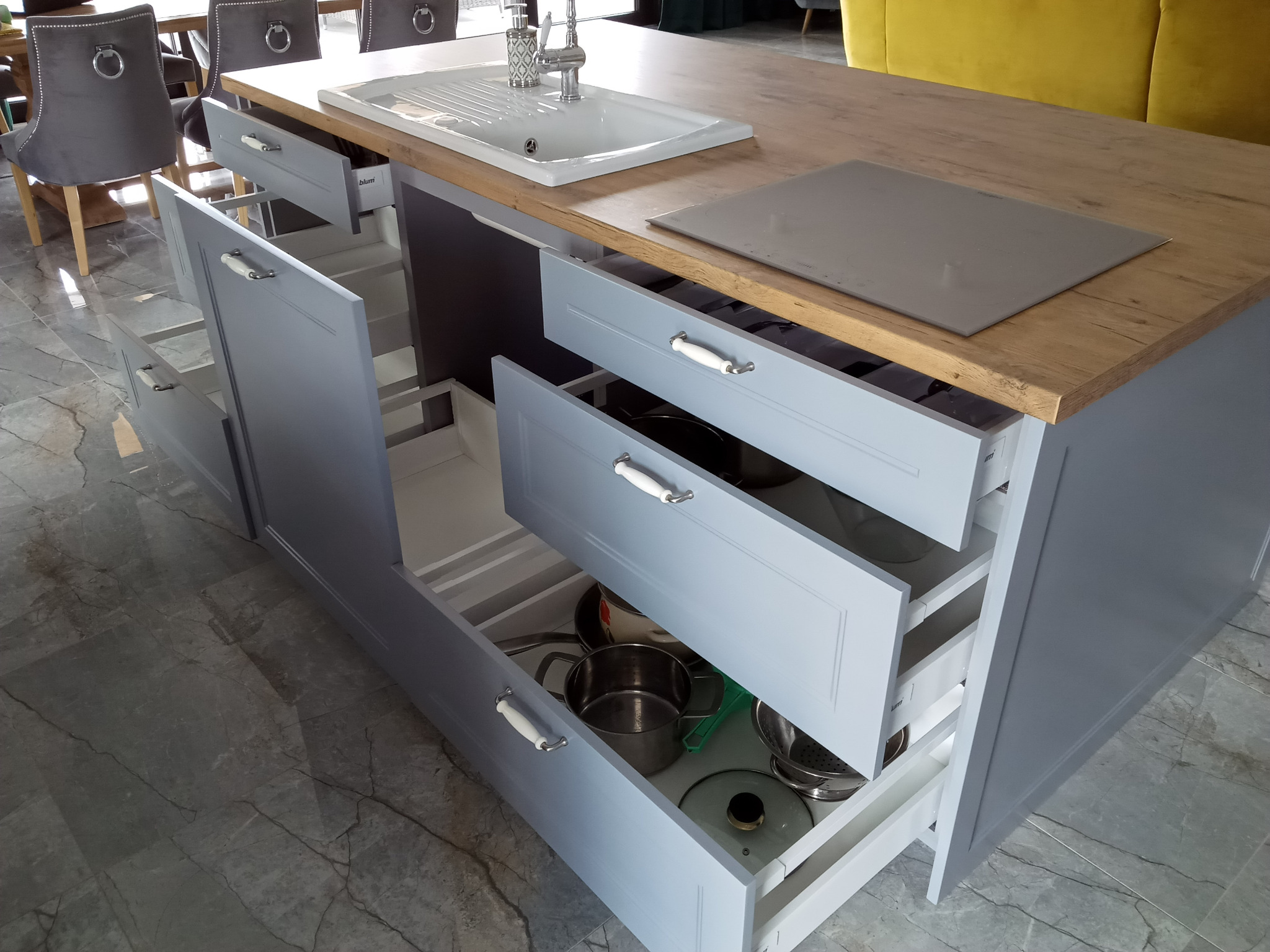 Wysuwane szuflady w szarej wyspie kuchennej z drewnianym blatem, z widocznymi naczyniami i garnkami w środku, umywalka i płyta indukcyjna na blacie, marmurowa podłoga.