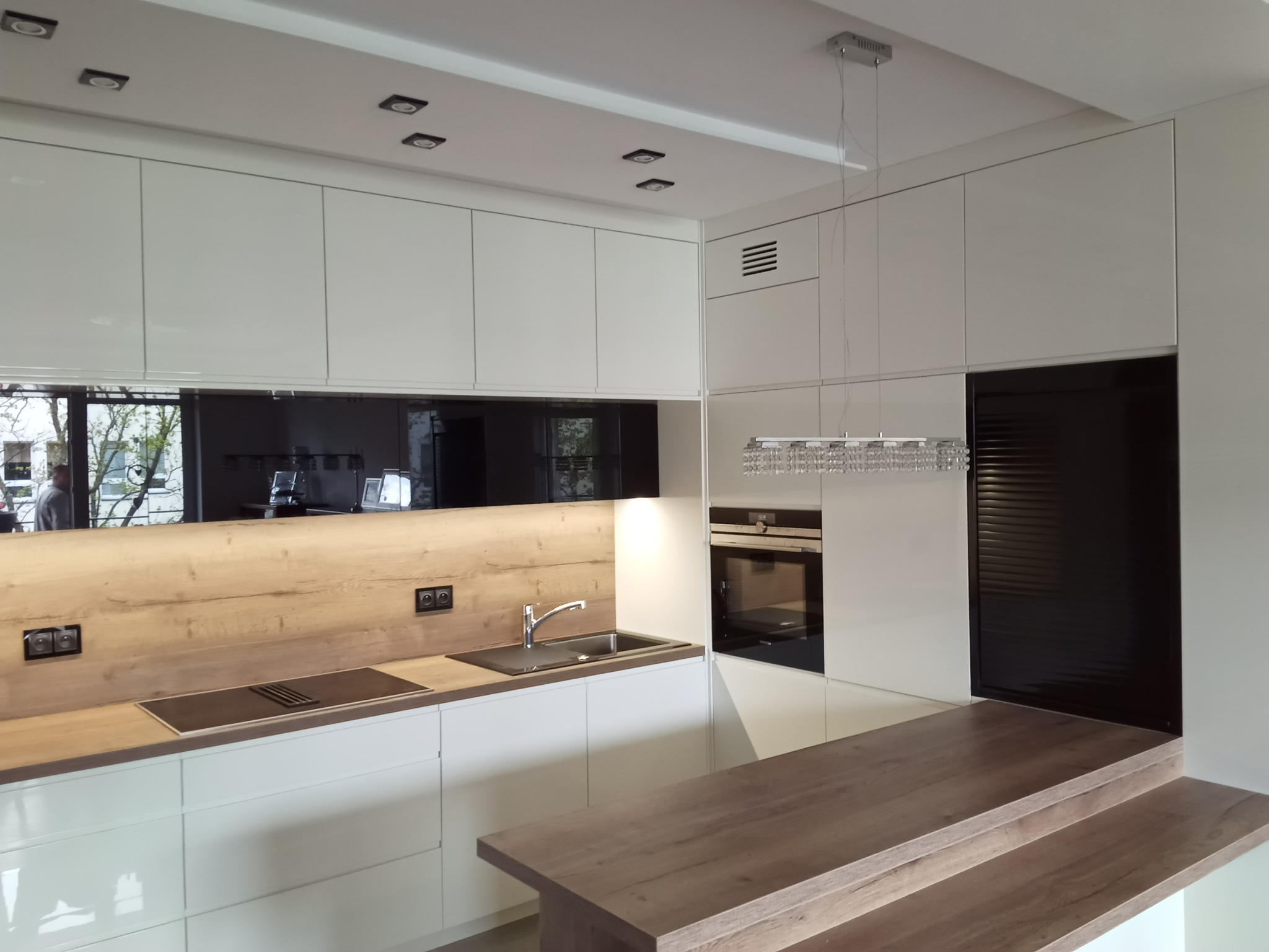 Nowoczesna kuchnia z białymi, lakierowanymi frontami szafek, drewnianym blatem i czarnym panelem nad blatem roboczym, oświetlona halogenami i wiszącą lampą kryształową.