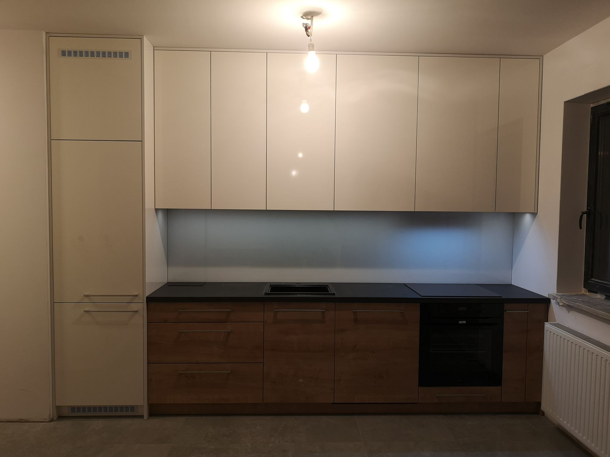 Jasna kuchnia z białymi szafkami górnymi i drewnianymi szafkami dolnymi z czarnym blatem, oświetlona pojedynczą żarówką.