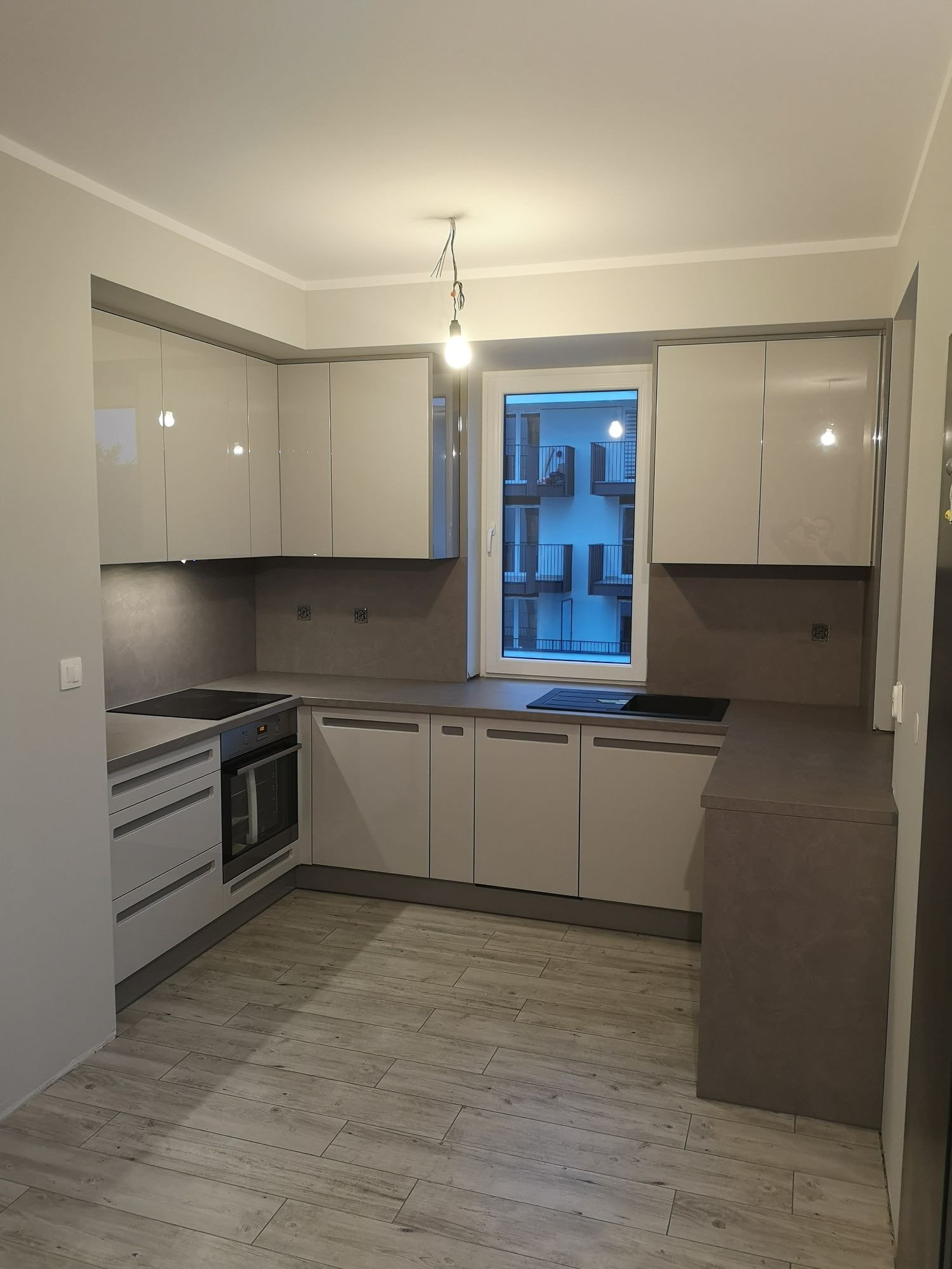 Nowoczesna kuchnia z błyszczącymi, kremowymi frontami szafek, szarym blatem i drewnopodobną podłogą; widok na budynek mieszkalny za oknem.