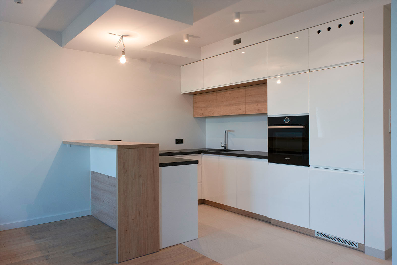 Nowoczesna kuchnia z białymi, lakierowanymi frontami, drewnianymi akcentami i czarnym blatem, oświetlona wiszącą żarówką i reflektorami sufitowymi, z widocznym barkiem.