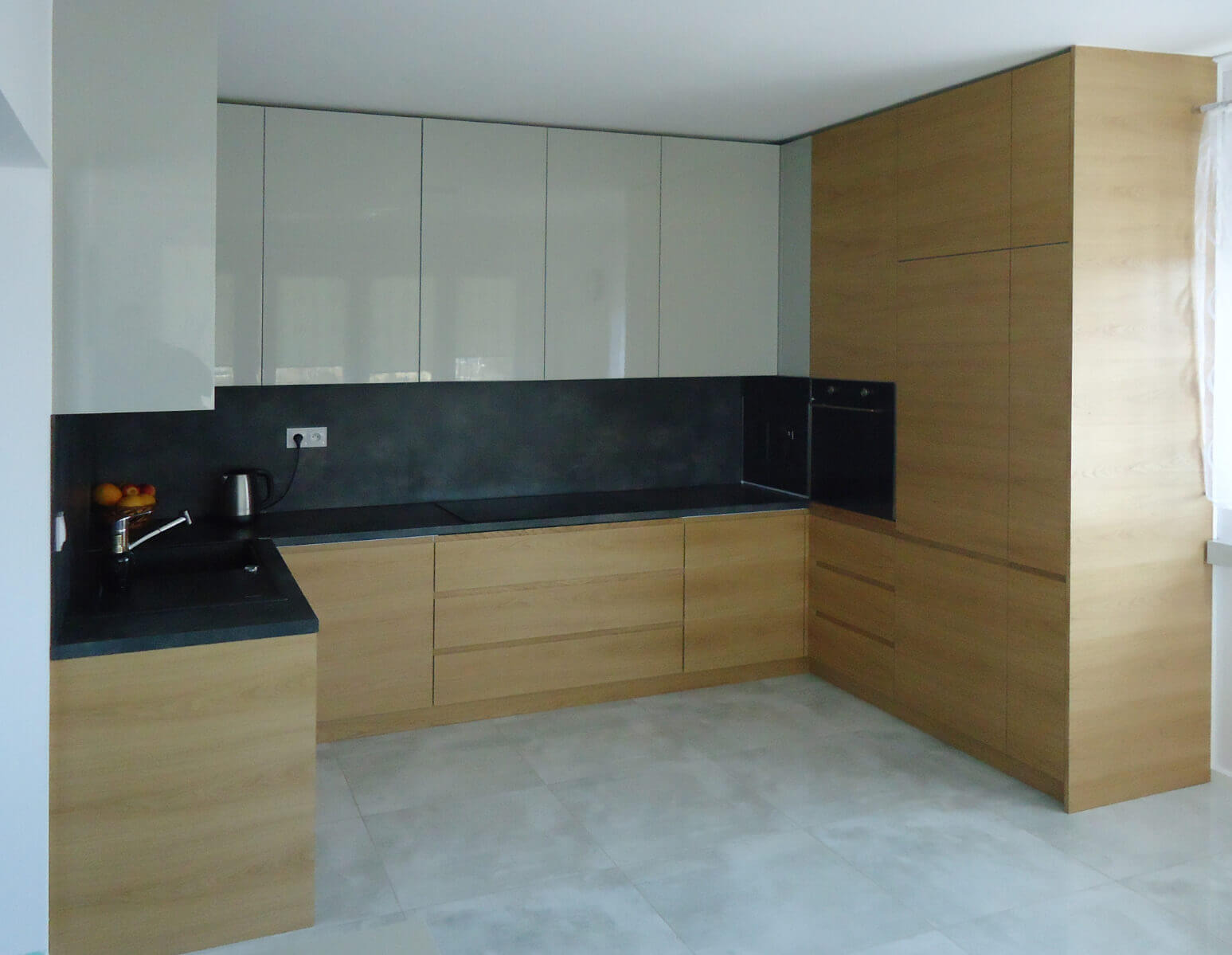 Nowoczesna kuchnia z frontami w dwóch kolorach: jasnoszarym połysku i drewnopodobnym, z czarnym blatem i ścianą nad blatem, widoczne owoce w misce przy zlewie