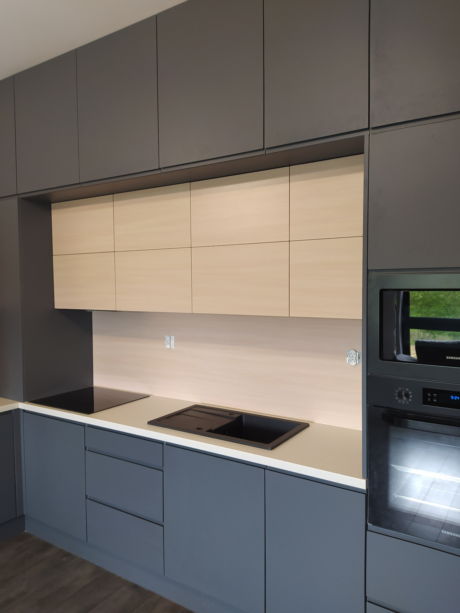Nowoczesna kuchnia w odcieniach szarości z jasnymi, drewnopodobnymi górnymi szafkami, czarnym zlewem i płytą indukcyjną, oraz wbudowanym piekarnikiem Samsung.