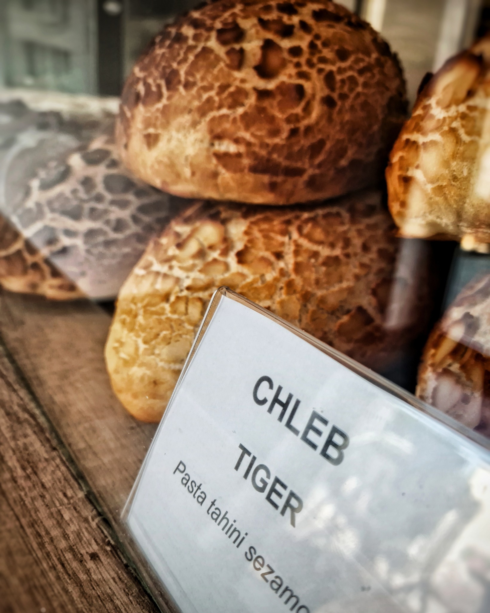 Tiger bread rolls stacked behind a plastic sign reading 'CHLEB TIGER Pasta tahini sezamo', showcasing the bread's distinctive cracked crust.