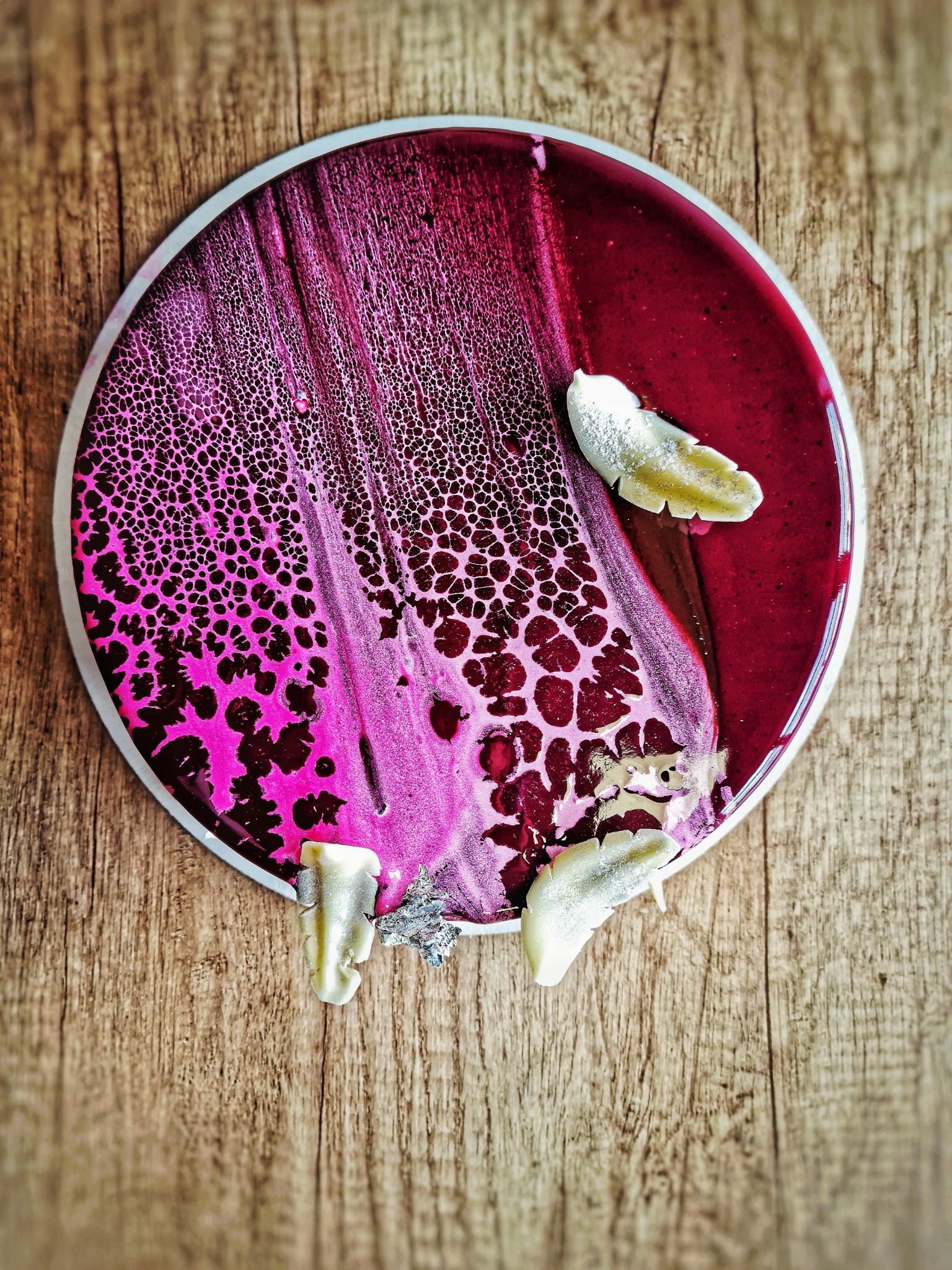 Abstrakcyjna kompozycja na drewnianym tle: okrągły talerz z różowo-bordową masą o strukturze siateczki i płatkami w srebrnym lukrze.