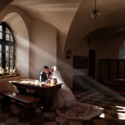 Para młoda siedzi przy drewnianym stole w gotyckim wnętrzu, oświetlona promieniami słońca wpadającymi przez okno z roślinami na parapecie, suknia panny młodej rozłożona na posadzce w szachownicę.