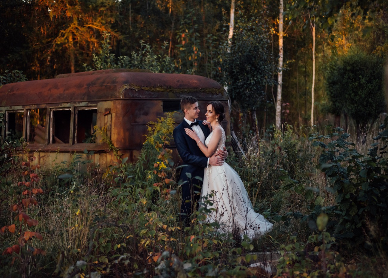 Para młoda w objęciach na tle zardzewiałego, opuszczonego autobusu, otoczona wysoką trawą i drzewami w ciepłym świetle zachodzącego słońca.