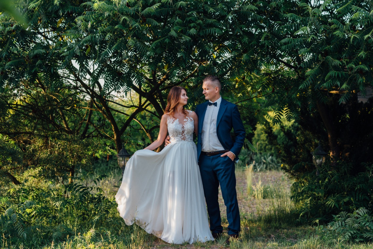 Para młoda w plenerze: Panna młoda w zwiewnej sukni z koronkową górą trzyma brzeg sukni, a pan młody w granatowym garniturze z muszką patrzy na nią z uśmiechem, stojąc pod rozłożystym drzewem.