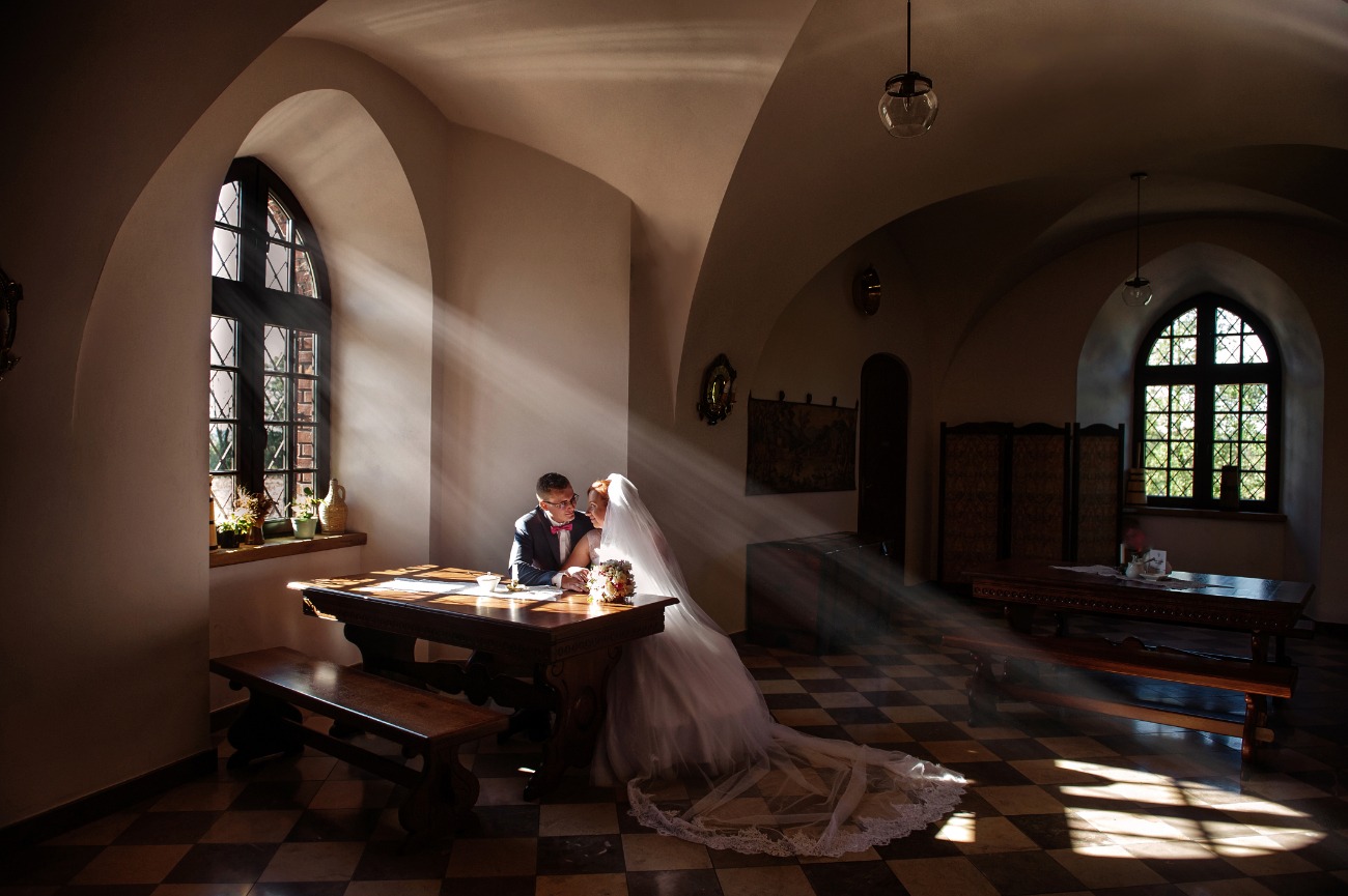 Para młoda siedzi przy drewnianym stole w gotyckim wnętrzu, oświetlona promieniami słońca wpadającymi przez okno z roślinami na parapecie, suknia panny młodej rozłożona na posadzce w szachownicę.