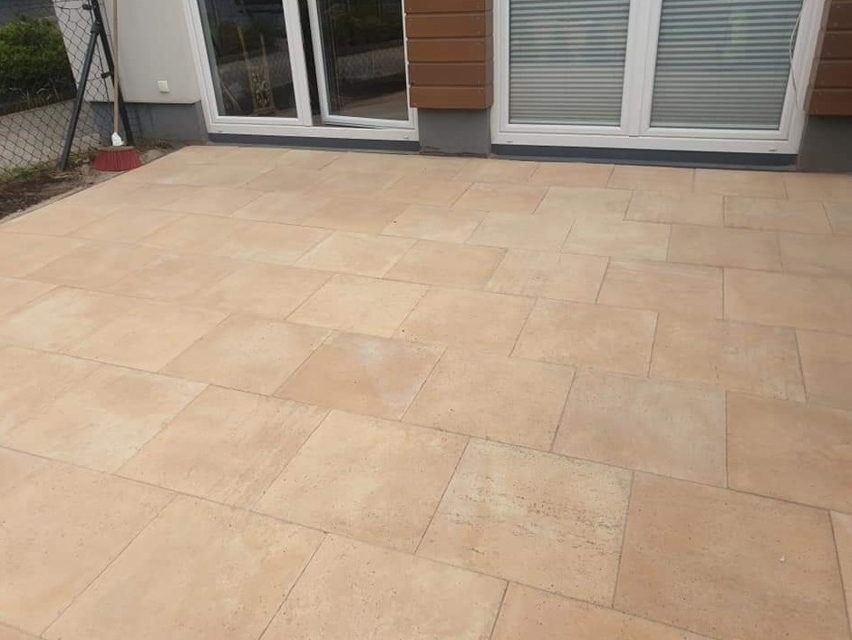 Ułożona kostka brukowa w odcieniach beżu tworząca taras przed budynkiem z białymi oknami i brązową elewacją, widoczny fragment ogrodzenia i miotła oparta o ścianę.