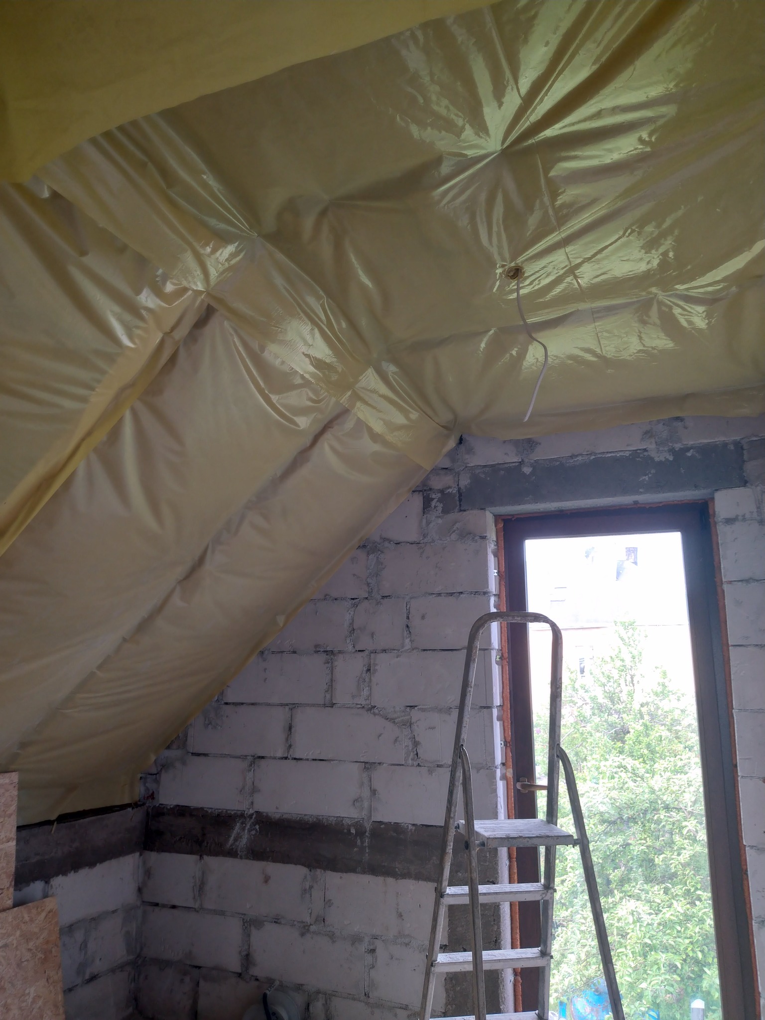 Poddasze w trakcie izolacji, widoczna żółta folia paroizolacyjna na skosach dachu, ściany z białych bloczków, drabina przystawiona do okna z widokiem na zieleń.
