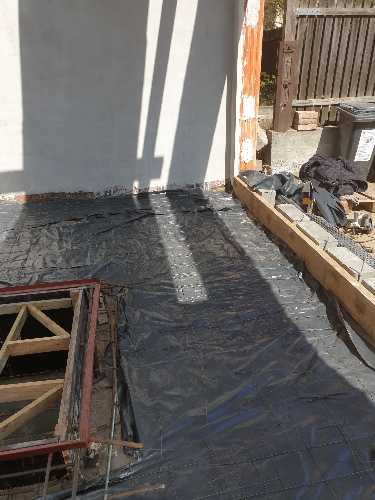 Przygotowanie podłoża pod fundament: czarna folia izolacyjna i siatka zbrojeniowa na gruncie, częściowo w drewnianym szalunku, z fragmentem ściany z cegły dziurawki w tle.