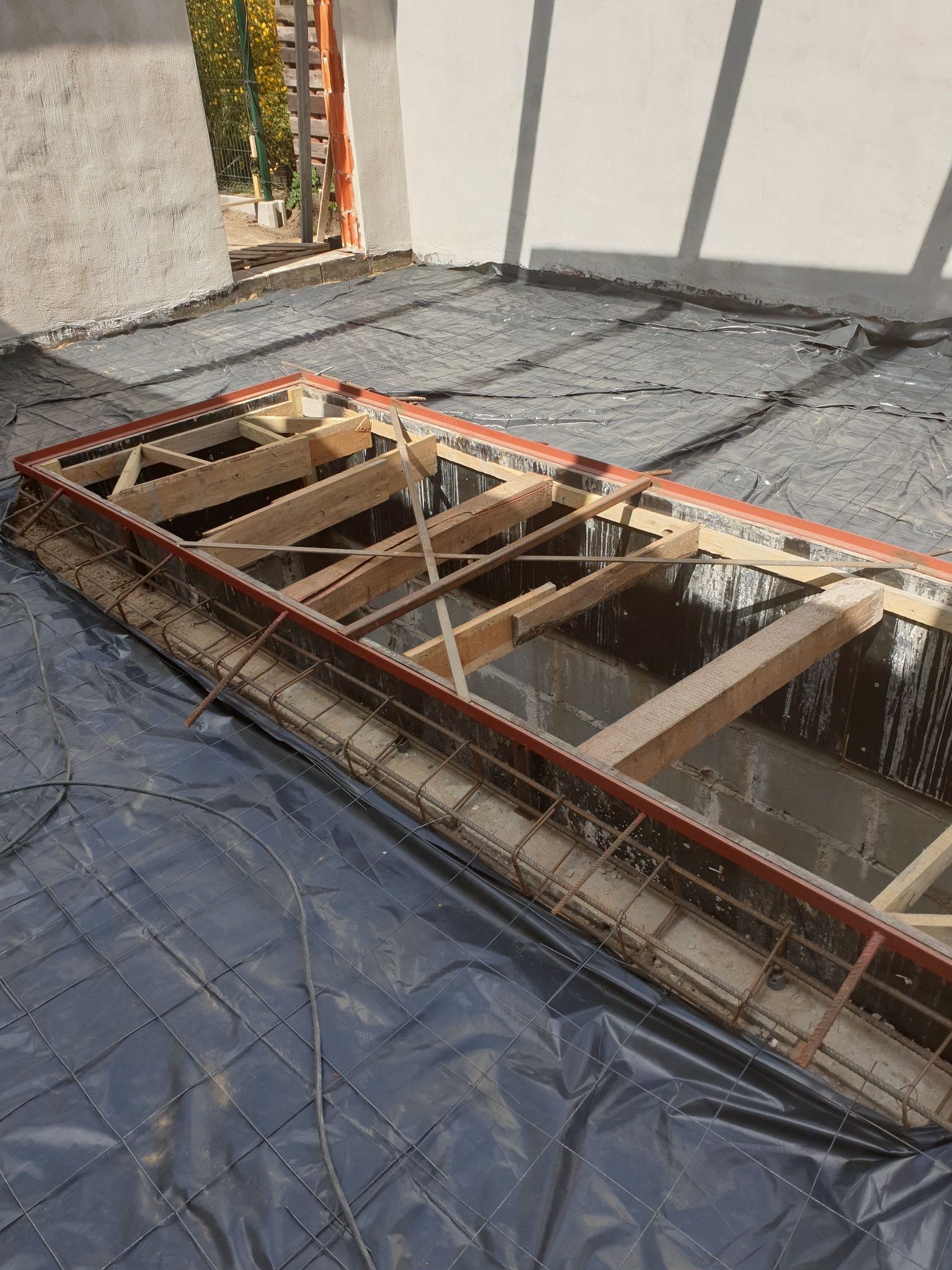 Zbrojenie w szalunku pod wylewkę fundamentową, widoczne drewniane podpory i czarna folia izolacyjna na podłożu.