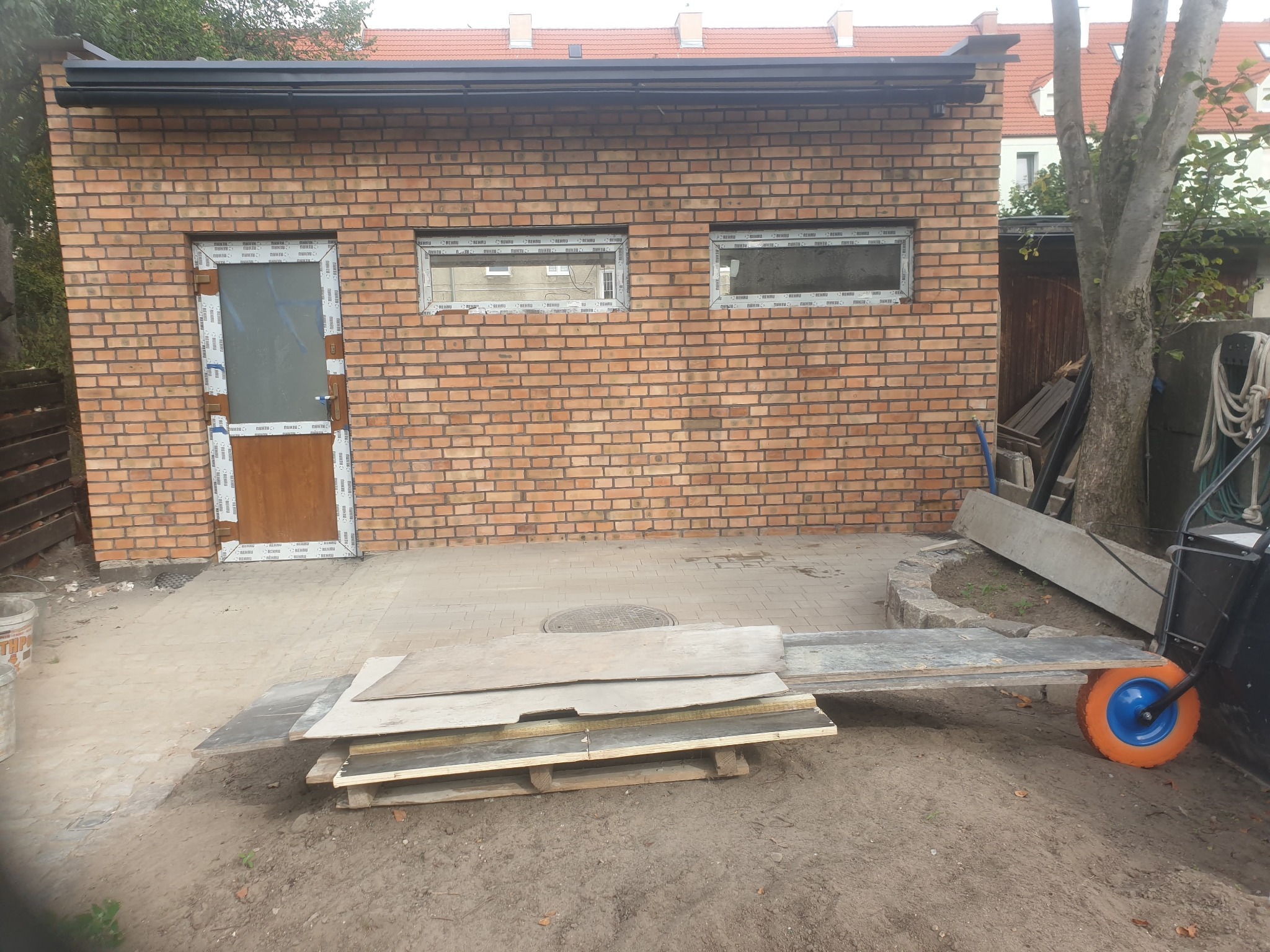 Murowany budynek gospodarczy z częściowo zamontowaną stolarką okienną i drzwiową, widoczne przygotowane płyty budowlane i wózek transportowy