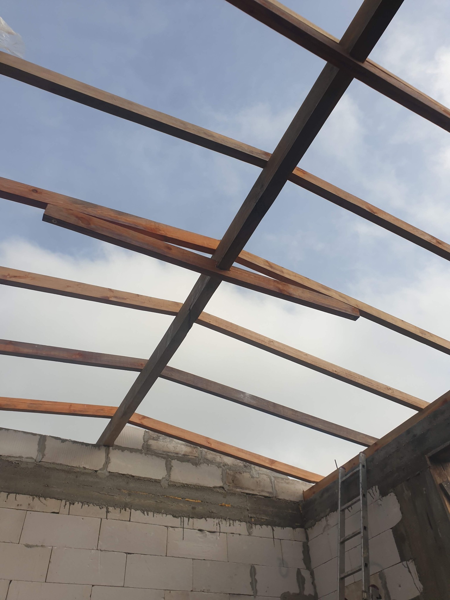 Wnętrze budynku w trakcie budowy, widok na drewnianą konstrukcję dachu z perspektywy dolnej, ściany z bloczków betonowych, drabina oparta o ścianę.
