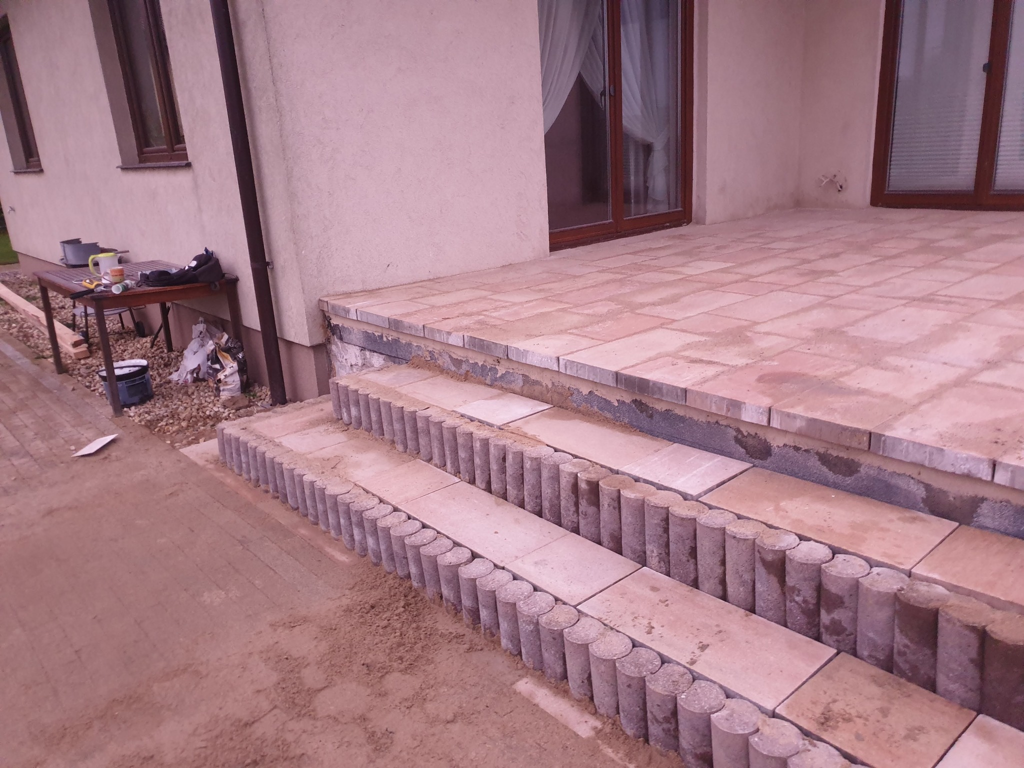 Wykończone schody wejściowe do domu, wyłożone płytami chodnikowymi i obrzeżami w kształcie walca, widoczne narzędzia i materiały budowlane na stole w tle.