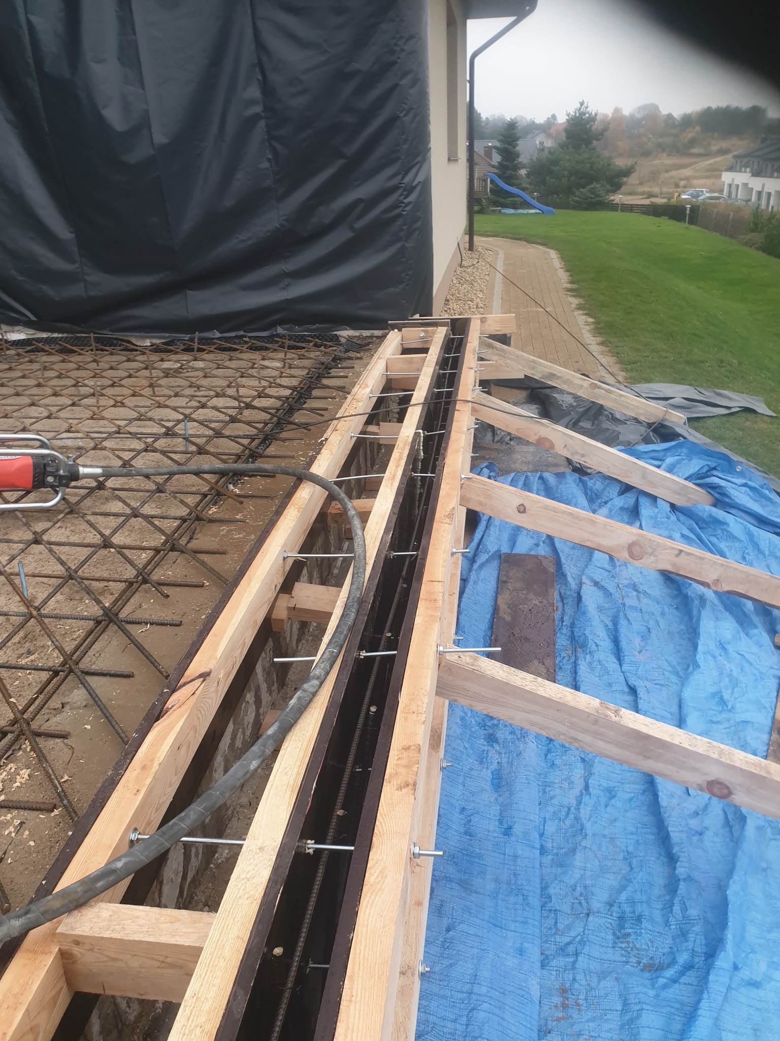 Zalewanie betonem zbrojonej konstrukcji szalunkowej pod fundament, w tle widoczny dom jednorodzinny i trawnik.