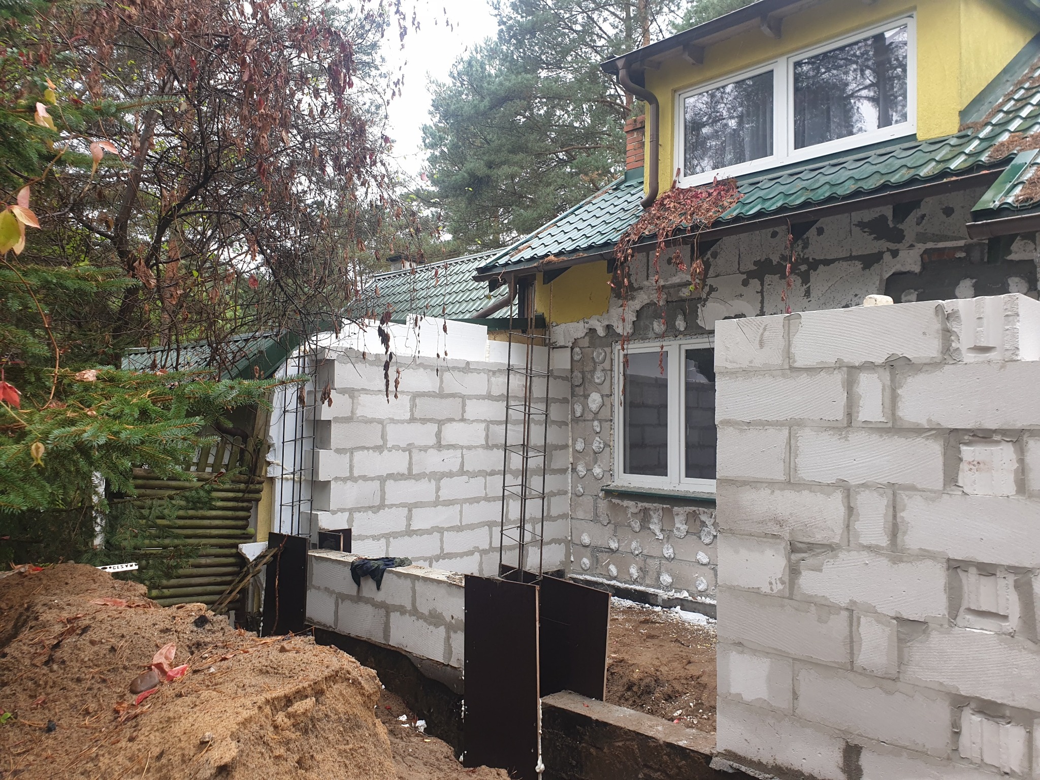 Wznoszenie ścian domu z bloczków betonowych, widoczny wykop pod fundamenty, szalunki i zbrojenie, w tle elewacja budynku z widocznymi śladami ocieplenia.